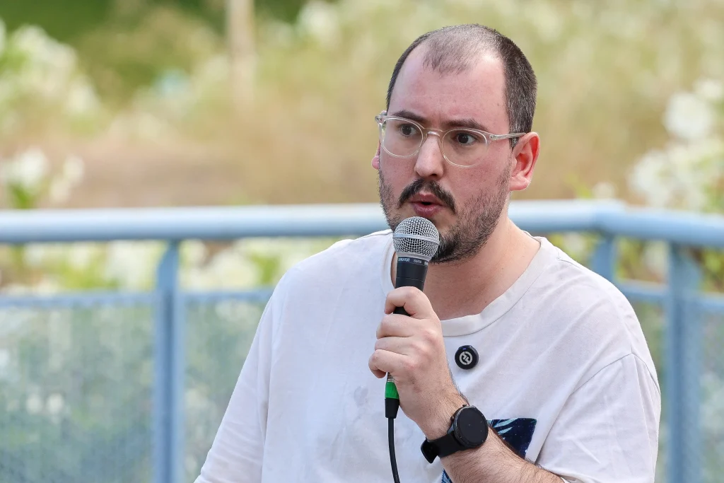 Una persona con el pelo corto, gafas y barba clara sostiene un micrófono mientras habla al aire libre en el evento «Política en la Calle» en Rivas. Lleva una camiseta blanca y un reloj de pulsera negro; detrás de ella se ve vegetación borrosa y una valla o barandilla azul.
