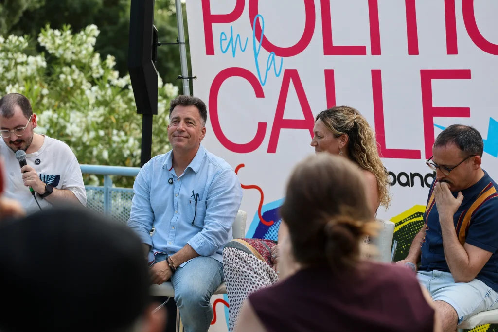 Cuatro personas forman un grupo al aire libre frente a un cartel en el que se lee «Política en la Calle». Pedro del Cura sostiene un micrófono mientras los demás escuchan y debaten sobre el modelo de ciudad de Rivas. En primer plano se ve al público.