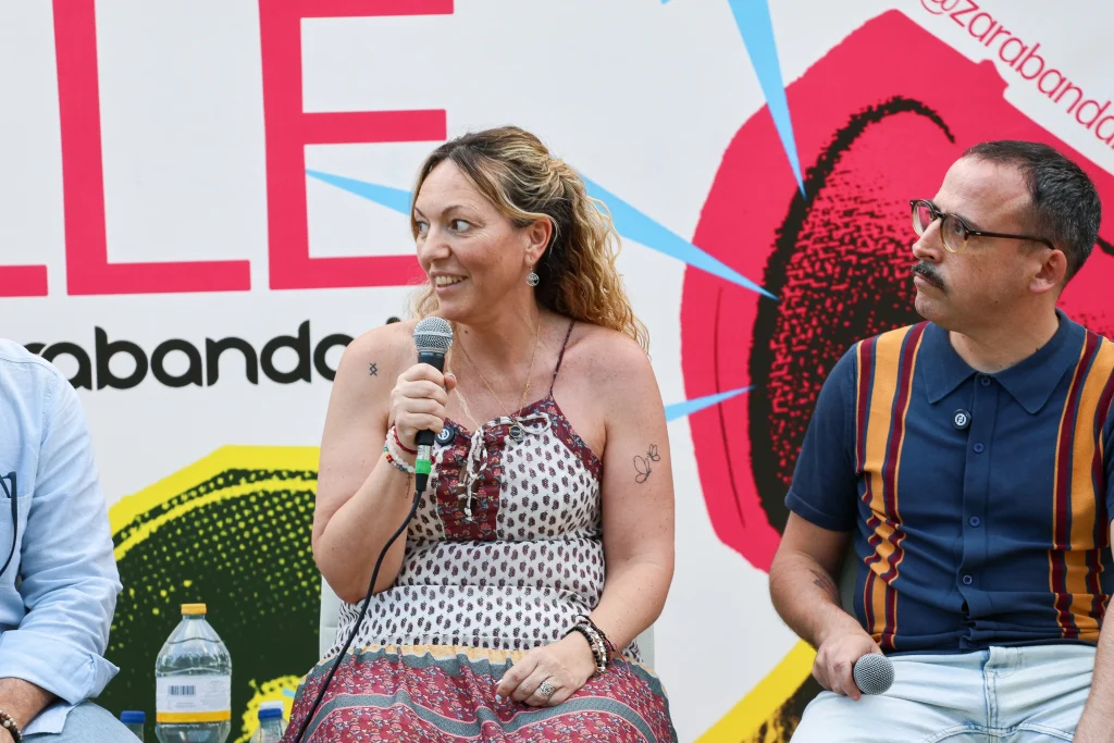 Una mujer con un vestido estampado sostiene un micrófono y sonríe mientras está sentada junto a Pedro del Cura, que lleva gafas y una camisa a rayas, durante el evento «Política en la Calle», celebrado al aire libre. Detrás de ellos se ve un colorido telón de fondo gráfico.