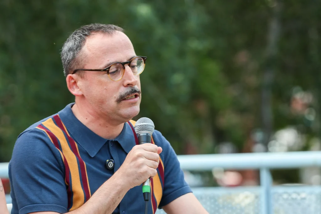 Una persona con el pelo corto, gafas y bigote sostiene un micrófono y habla al aire libre en el evento «Política en la Calle». Viste una camisa azul con rayas verticales de varios colores y se encuentra de pie entre la vegetación en Rivas, posiblemente en Pedro del Cura.