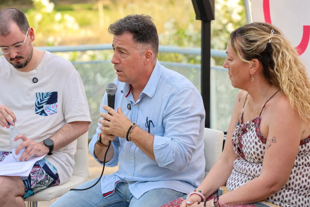 Tres personas están sentadas al aire libre en la ciudad de Rivas. El hombre del centro, posiblemente Pedro del Cura, habla por un micrófono durante el programa «Política en la Calle», mientras los demás escuchan y revisan unos papeles rodeados de vegetación y con un cartel parcialmente visible.