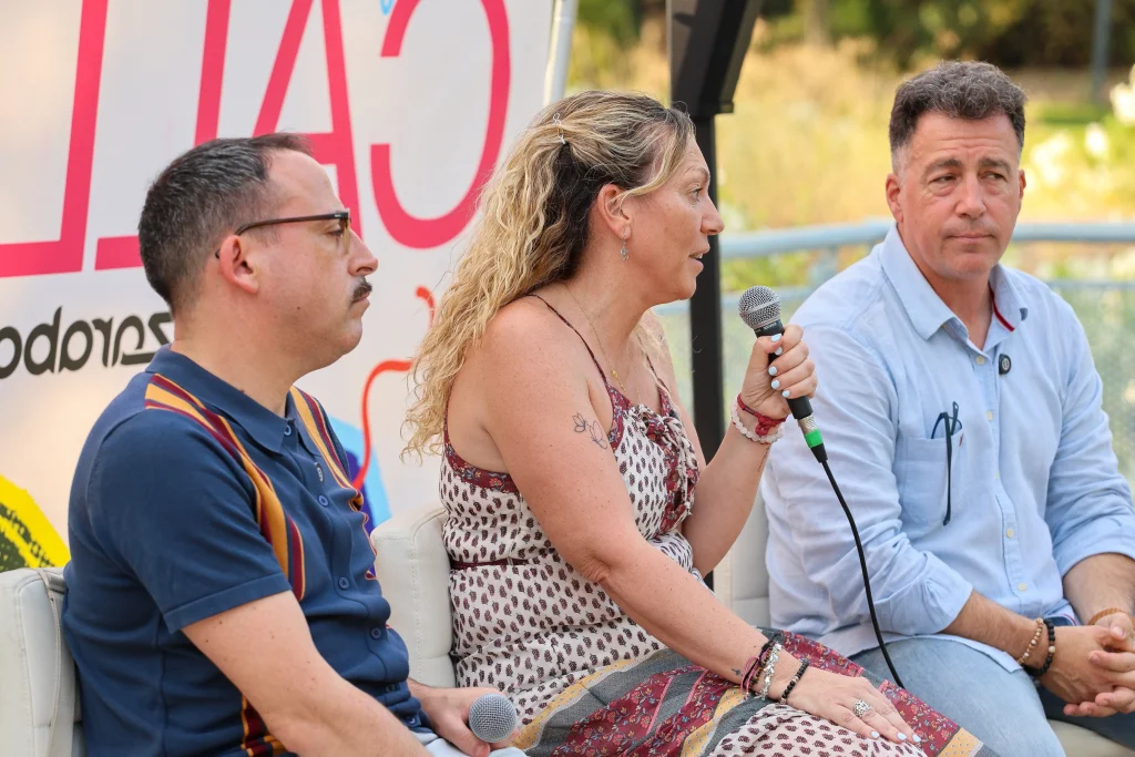Tres personas están sentadas en sillas al aire libre, conversando. La mujer del centro sostiene un micrófono —probablemente durante el evento «Política en la Calle»—, mientras Pedro del Cura y otro hombre escuchan con atención. Detrás de ellos se ve parcialmente un cartel con letras rojas grandes.