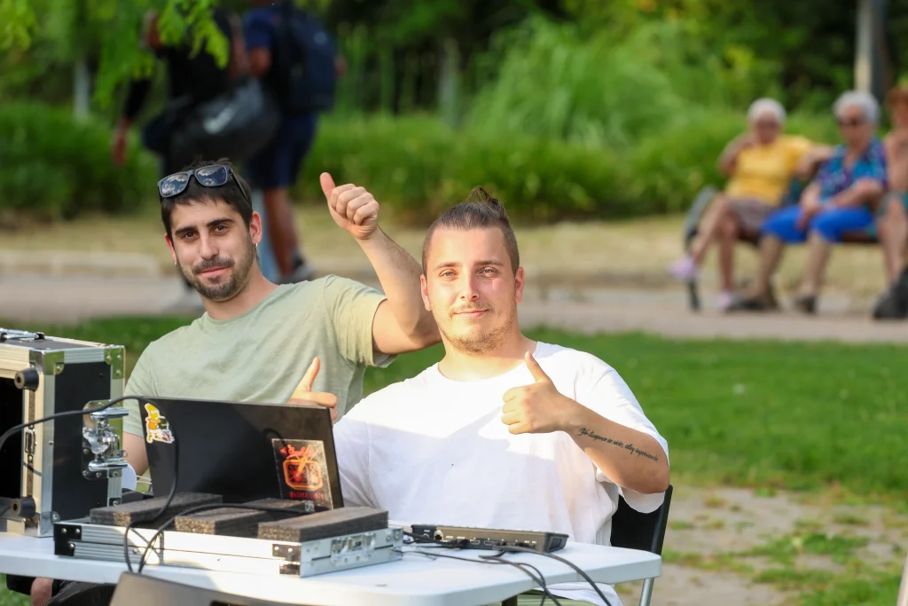 Dos personas, posiblemente Pedro del Cura y Vanessa Millán, están sentadas al aire libre en una mesa con aparatos electrónicos y un ordenador portátil, haciendo un gesto de aprobación con el pulgar. Al fondo se ve césped, árboles y gente en los bancos, lo que refleja el modelo de ciudad de Rivas.