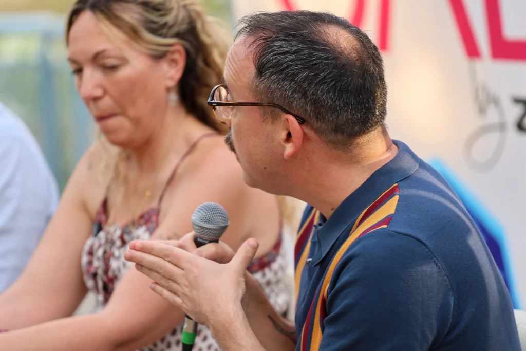 Un hombre que sostiene un micrófono habla mientras gesticula con las manos; una mujer está sentada a su lado con expresión impasible. Ambos se encuentran frente a un cartel de fondo colorido en el evento «Política en la Calle», debatiendo sobre el modelo de ciudad de Rivas.