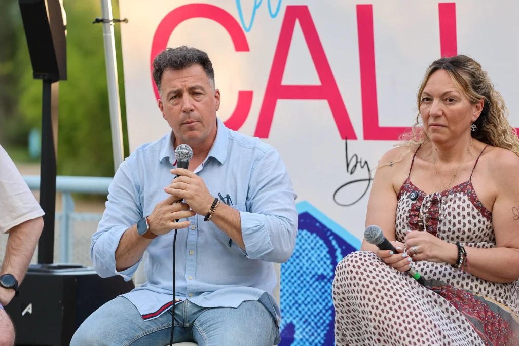 Un hombre con una camisa azul claro y una mujer con un vestido sin mangas están sentados uno al lado del otro, cada uno con un micrófono en la mano, frente a un cartel de «Política en la Calle», debatiendo sobre el modelo de ciudad de Rivas.