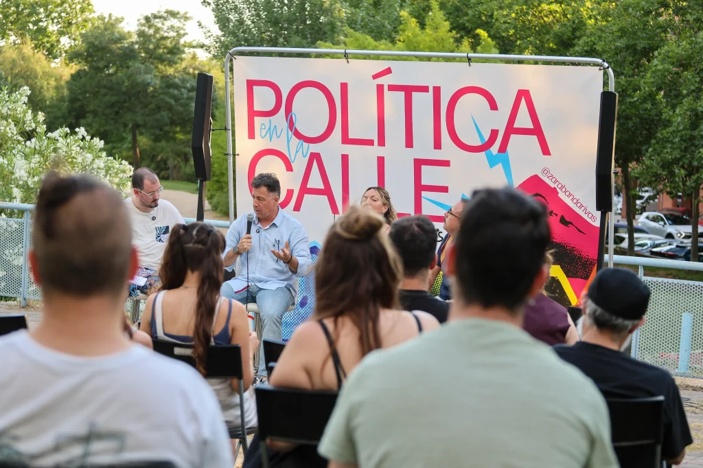 Un grupo de personas está sentado al aire libre en Rivas, frente a un panel de ponentes —Pedro del Cura y Vanessa Millán— situado delante de un gran cartel en el que se lee «Política en la calle». Al fondo se ven árboles y coches aparcados.