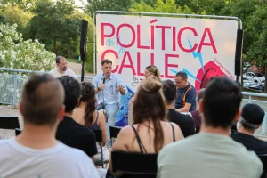 Cuatro personas forman parte de una mesa redonda frente a un público al aire libre, hablando bajo un cartel que reza «POLÍTICA en la CALLE». Entre ellas se encuentran Vanessa Millán y Pedro del Cura, que debaten sobre el modelo de ciudad de Rivas con árboles y una valla al fondo.