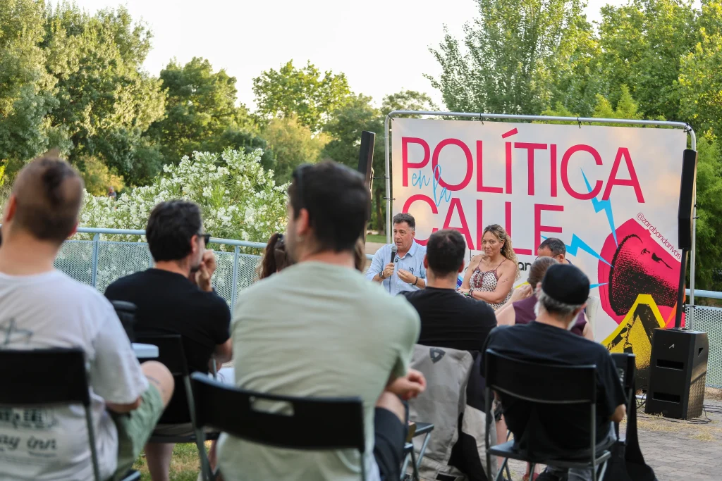 Un grupo de personas está sentado al aire libre frente a un panel de tres ponentes, entre los que se encuentran Vanessa Millán y Pedro del Cura, delante de un gran cartel en el que se lee «Política en la Calle». Los árboles y la vegetación enmarcan el debate sobre el modelo de ciudad de Rivas.