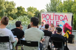 Un grupo de personas está sentado al aire libre frente a un panel de tres ponentes, entre los que se encuentran Vanessa Millán y Pedro del Cura, delante de un gran cartel en el que se lee «Política en la Calle». Los árboles y la vegetación enmarcan el debate sobre el modelo de ciudad de Rivas.