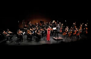 Una orquesta sinfónica actúa en el escenario con músicos que tocan instrumentos de cuerda, viento-madera, metal y percusión. Una solista vestida de rojo está de pie cerca del director, mientras un borrador automático de la iluminación crea un dramático fondo oscuro.