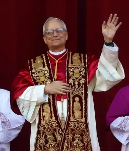 Un clérigo de la Iglesia Católica, ataviado con ornamentadas vestiduras religiosas, permanece de pie delante de una cortina roja, sonriendo y levantando la mano derecha en un gesto, con otras personas parcialmente visibles a ambos lados.