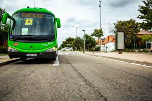 Un autobús escolar verde con la inscripción «EN BUS AL COLE», que forma parte de las rutas escolares 2026/2027, está aparcado en una parada de autobús en una calle de las afueras. Las casas, los árboles, la acera y un cielo nublado conforman el fondo.