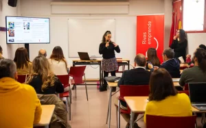 Una mujer está de pie al frente del aula hablando por un micrófono, con una presentación —titulada «Borrador automático»— proyectada en la pantalla que hay detrás de ella. Los participantes, sentados, miran hacia delante y escuchan. A la derecha, una pancarta roja reza «Rivas emprende».