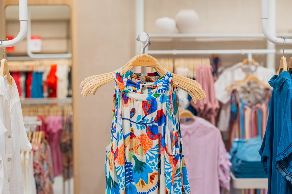 Un vestido sin mangas con un colorido estampado floral cuelga de una percha de madera en la tienda «Encuentro Modas» del Centro Comercial H2O, en Madrid, rodeado de otras prendas colgadas en percheros y estanterías al fondo.
