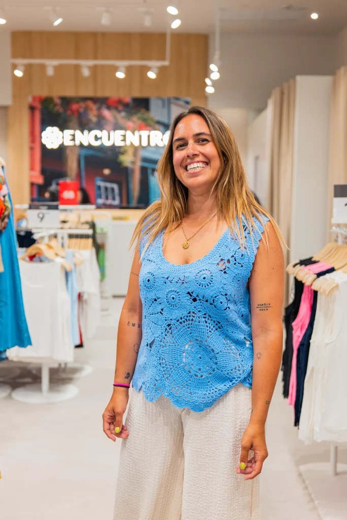 Una mujer de pelo largo, vestida con un top azul de ganchillo y pantalones blancos, posa sonriente en el interior de la luminosa tienda «Encuentro Modas» del Centro Comercial H2O de Madrid, con percheros llenos de ropa y un cartel que dice «ENCUENTRO» al fondo.