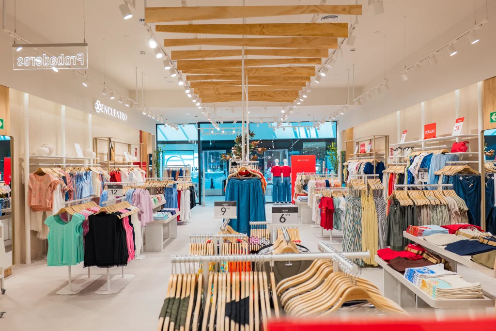 El interior de una tienda de ropa muy bien iluminada en el Centro Comercial H2O de Madrid, con percheros ordenados en los que se exponen las coloridas prendas de Encuentro Modas, etiquetas con los precios y un techo decorativo de madera. Los grandes ventanales de la parte trasera dejan entrar la luz natural.