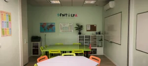 Un aula en Rivas con paredes de color verde claro, sillas de colores alrededor de las mesas, un mapamundi, la tabla periódica, una pizarra blanca, estanterías y una pequeña planta. En la pared se lee «Pentalfa Centro de Estudios», ideal para clases de refuerzo en verano.