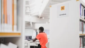 Una persona que lleva una camiseta naranja está sentada ante un escritorio en una luminosa biblioteca de Madrid, trabajando con el ordenador. En primer plano se ven estanterías y un cartel con la inscripción «Narrativa», lo que resalta el animado ambiente de las bibliotecas de Madrid en verano.