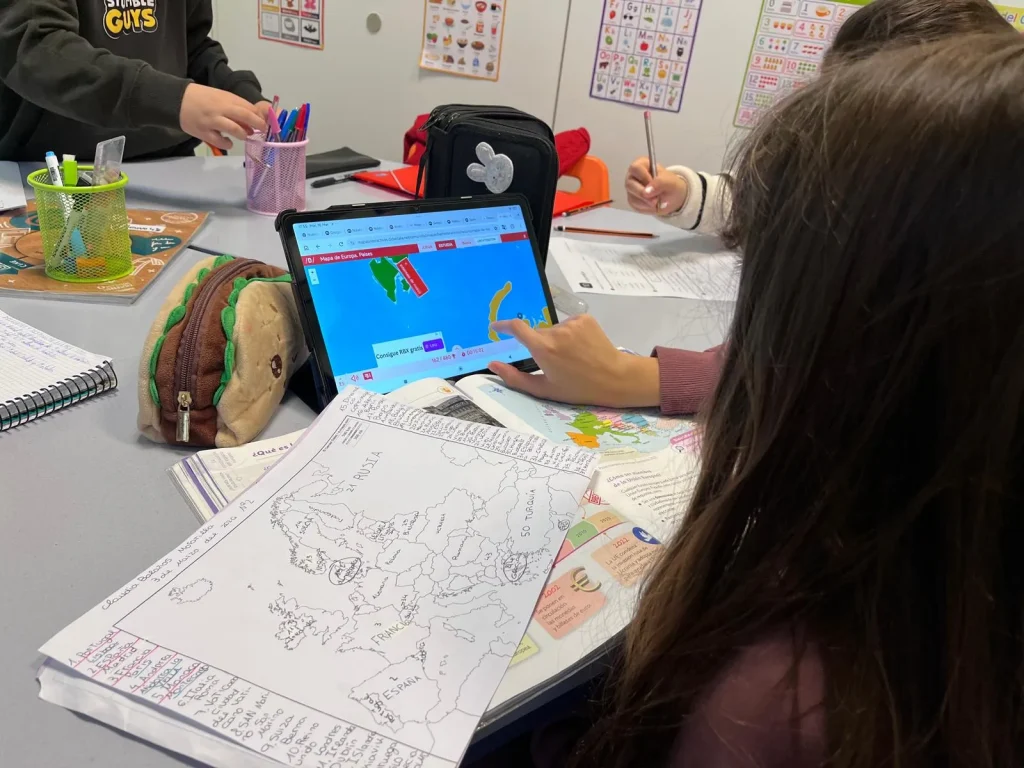 Un alumno de Rivas asiste a clases de refuerzo, estudiando en un pupitre con un mapa abierto y apuntes, mientras utiliza una tableta en la que se ve un mapa. Otros alumnos escriben y tienen material escolar, como bolígrafos y estuches, esparcido por la mesa.