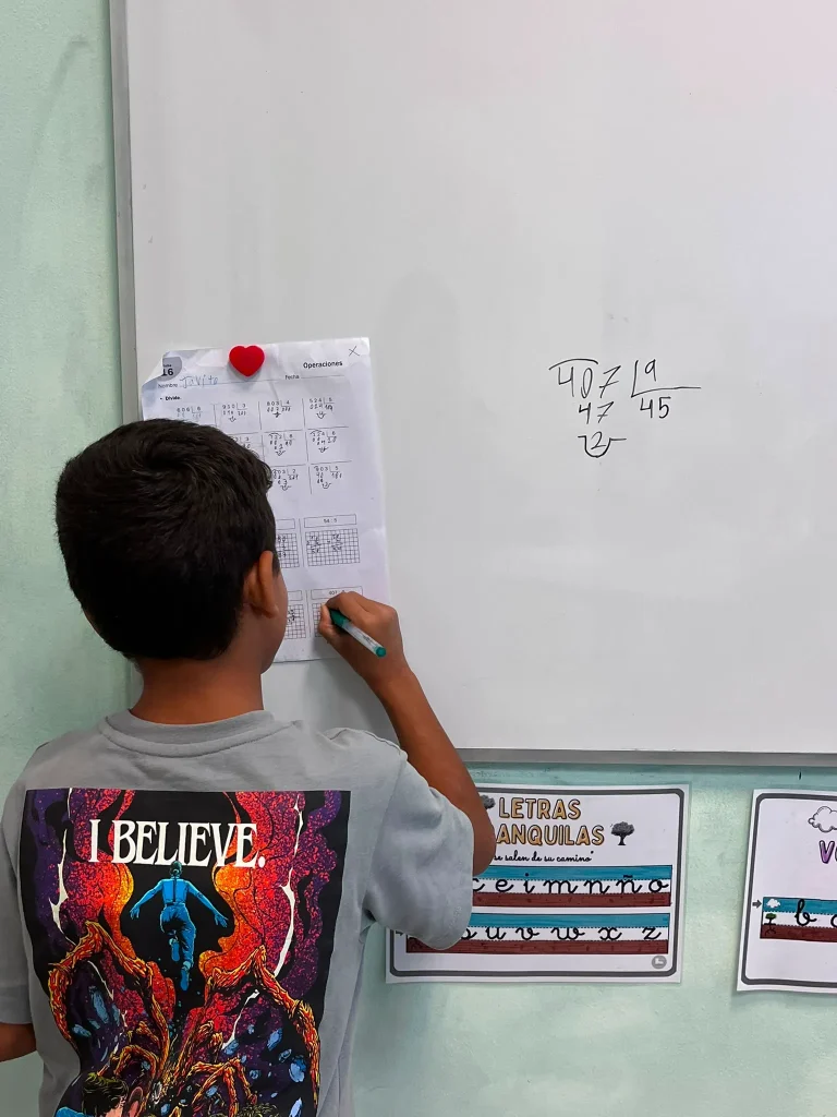Un niño asiste a clases de refuerzo en Rivas, de pie junto a una pizarra blanca para resolver un problema de división. Sostiene un rotulador y consulta su hoja de ejercicios, mientras que en su camiseta se ve un dibujo colorido con el texto «I BELIEVE».