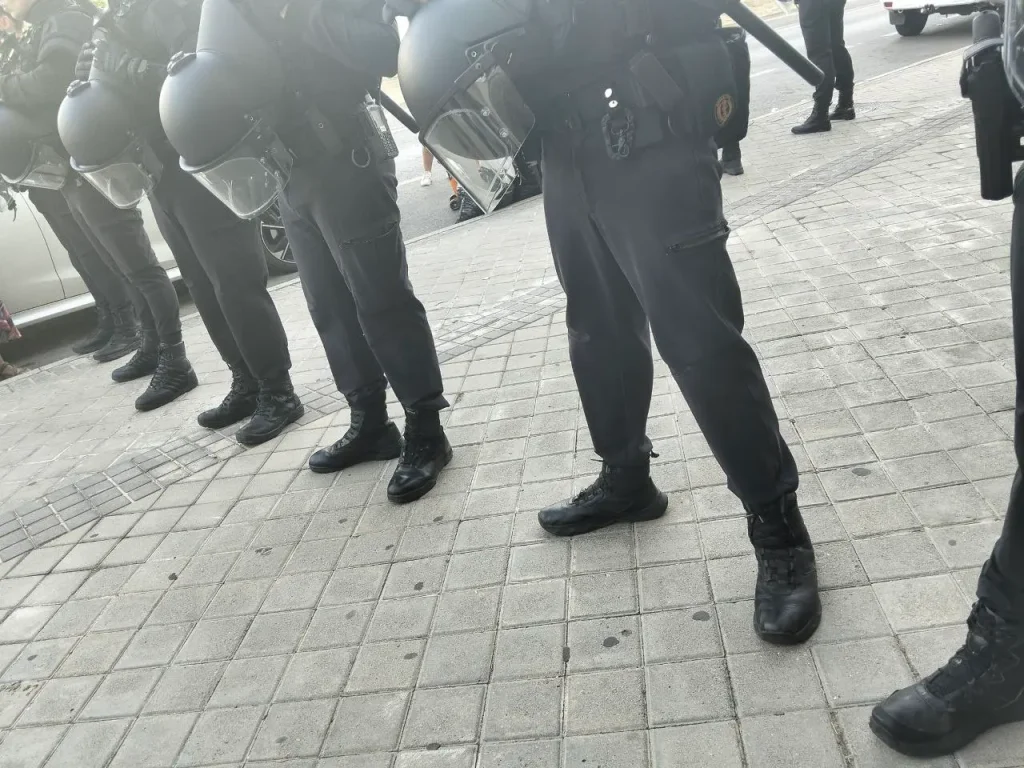 Una fila de agentes de policía, vestidos con equipo antidisturbios negro y cascos, se encuentra de pie en una acera de baldosas, de espaldas a la cámara, durante un desalojo judicial cerca de las viviendas EMV. No se les ve la cara; solo se ven la parte inferior de sus cuerpos y su equipo.