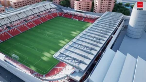Vista aérea del Estadio de Vallecas, en la que se aprecian los asientos rojos y blancos, el campo verde, las gradas parcialmente cubiertas, los edificios altos cercanos y el logotipo de la Comunidad de Madrid en la esquina superior derecha, en medio de las obras de reforma en curso.