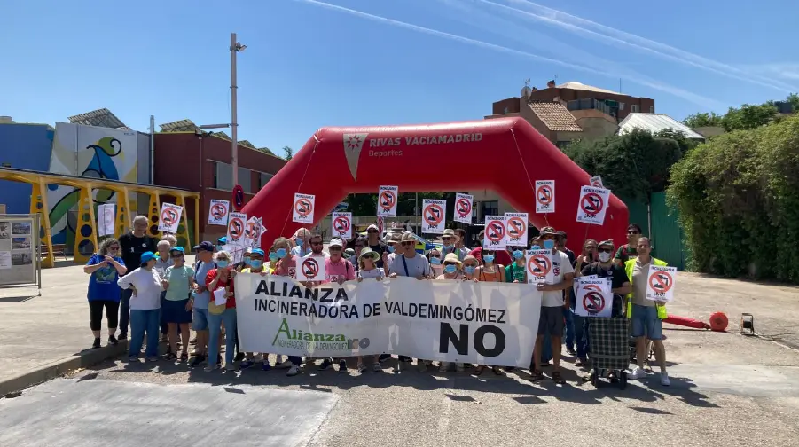 Un grupo de personas, entre ellas niños y adultos, permanecen al aire libre en Rivas sosteniendo carteles de "No" y una pancarta en la que se lee "Alianza Incineradora de Valdemingómez NO", apoyando el cierre de Valdemingómez frente a una estructura hinchable roja.