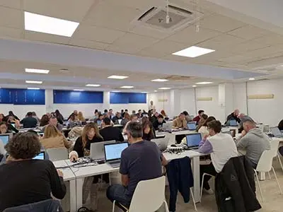 Una amplia sala llena de gente sentada en mesas, trabajando con sus ordenadores portátiles para Rivamadrid. El espacio tiene paredes blancas, un techo blanco con luces y persianas azules. El ambiente es de concentración y ajetreo, ideal para quienes buscan empleo o consultan la bolsa de empleo.