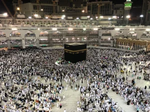 Miles de personas se reúnen en torno a la Kaaba de la Gran Mezquita de La Meca, vestidas en su mayoría de blanco, para peregrinar a La Meca. La mezquita está brillantemente iluminada y rodeada de arcos de varios pisos. Crece la preocupación por el golpe de calor debido al cambio climático.