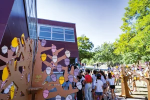 Un grupo de personas, entre las que hay niños, se reúne frente a un edificio moderno, cerca de unos árboles de cartón decorados con bellotas de papel de colores, en uno de los colegios de Rivas que participan en el proyecto 50/50 de ahorro de energía. La escena, luminosa y soleada, transmite un ambiente de comunidad y expectación ante el curso 2024/2025.