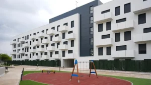 Un moderno edificio de apartamentos blanco con contraventanas negras, construido en el marco del Plan Estatal de Vivienda, se alza detrás de un pequeño parque infantil con columpios y juegos elásticos sobre un suelo de caucho rojo. El cielo está nublado.