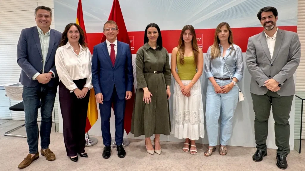 Siete personas están de pie en fila, posando para una foto de grupo en un interior de Madrid. Detrás de ellas hay dos banderas y un telón de fondo rojo y blanco con texto y logotipos, en homenaje a los logros de la PAU y a la «mejor nota». El grupo está formado por cuatro mujeres y tres hombres, todos ellos vestidos de forma elegante.
