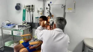 Un médico examina el oído de un niño con un instrumento médico en la consulta de una clínica, lo que pone de relieve la importancia de la salud de los menores. En el fondo se ven equipos médicos, material sanitario y un reloj de pared.