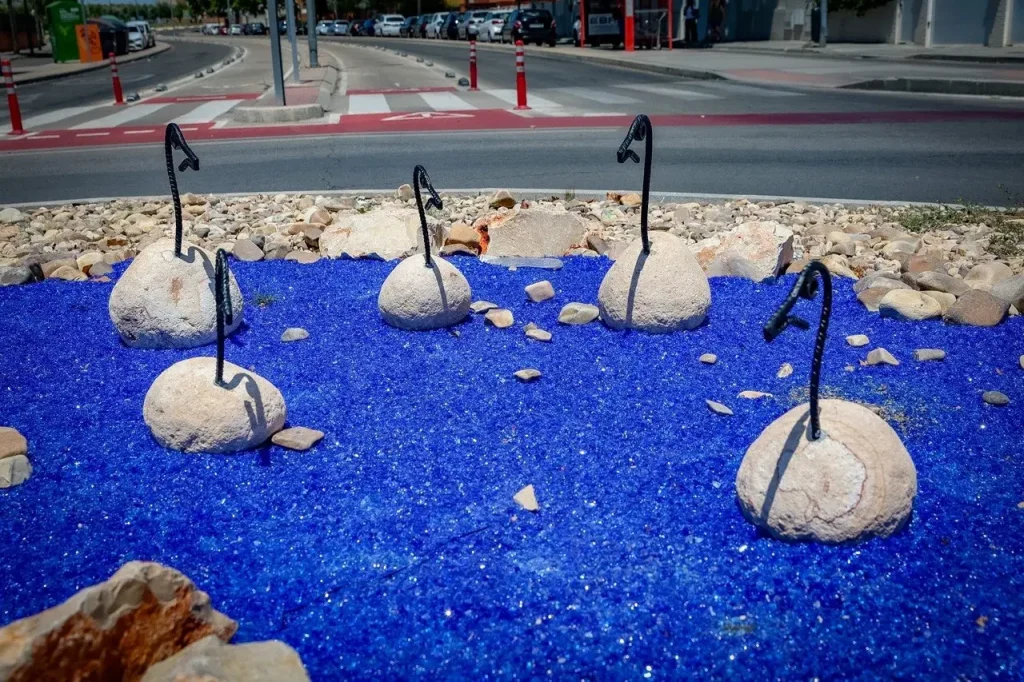 Una rotonda cuenta con grava azul mezclada con grandes rocas y varillas metálicas negras con forma de cuellos y cabezas de cisne, dispuestas de tal manera que recuerdan a cisnes en el agua. Situada en la avenida Pilar Miró, la instalación es un ejemplo llamativo de un proyecto vecinal que embellece el paisaje local. Al fondo se divisan una carretera y coches aparcados.