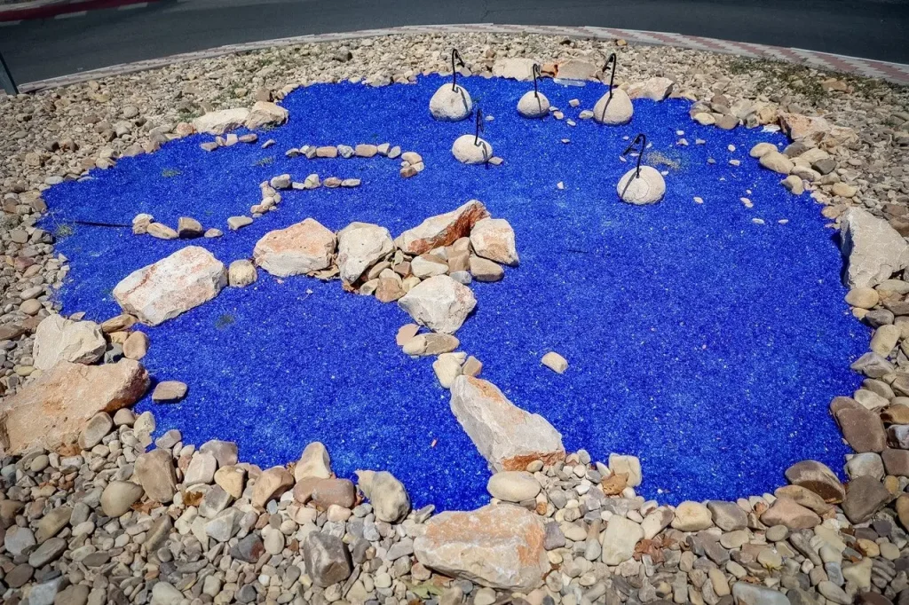 Un elemento paisajístico seco, compuesto por grava azul con forma de estanque y rodeado de rocas, embellece una de las rotondas de la avenida Pilar Miró, en Rivas. Varias rocas de mayor tamaño, con varillas metálicas, parecen pájaros estilizados posados en el agua.