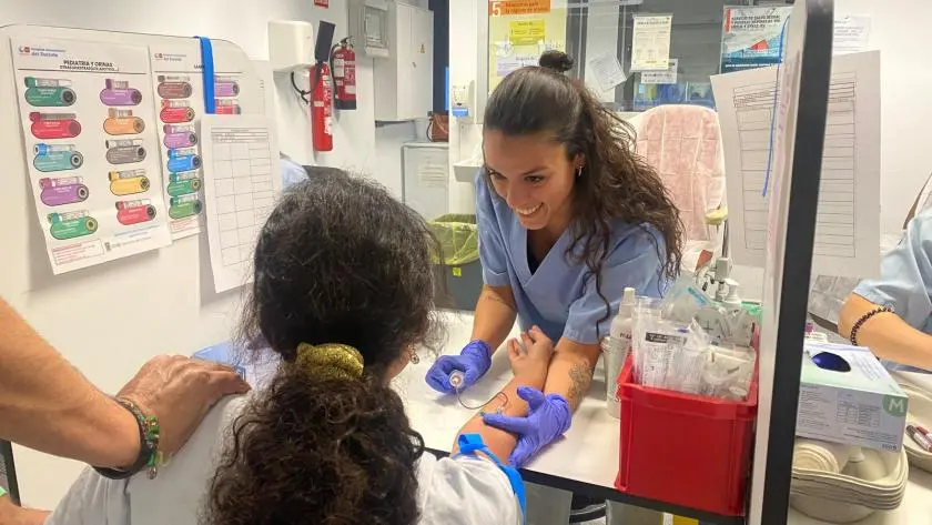 Una enfermera sonríe mientras extrae sangre a un paciente sentado en una consulta médica del Hospital del Sureste. El brazo del paciente descansa sobre la mesa y otra persona, de pie junto a él, le toca el hombro. Sobre la mesa se ven materiales sanitarios.