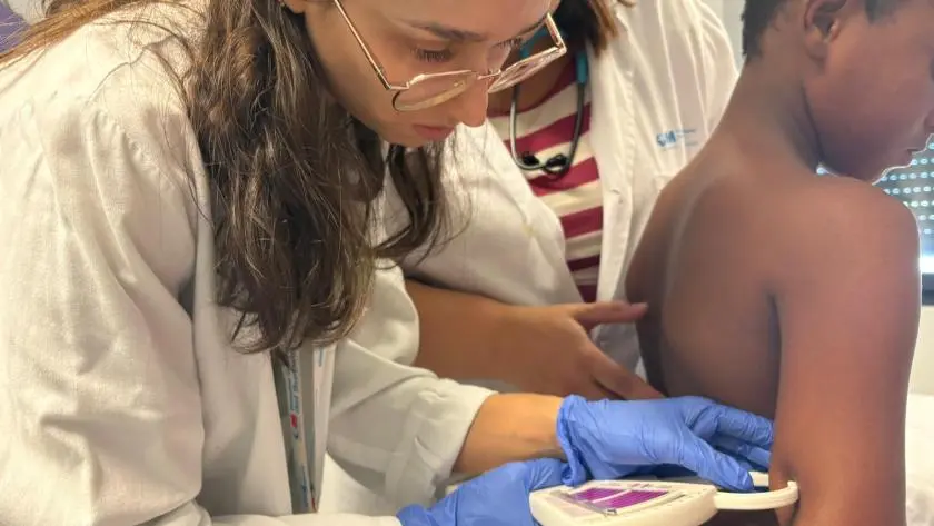 Un profesional sanitario, con bata blanca y guantes azules, utiliza un dispositivo médico en la parte superior del brazo de un niño, contribuyendo así a la salud de los menores. Otra persona, también con bata blanca, se encuentra cerca, colaborando en el procedimiento. El entorno parece clínico o médico, lo que refleja la calidad de la atención que se presta en el Hospital del Sureste.