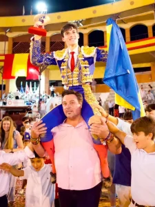 En el Circuito de Madrid 2026, una persona vestida con un traje ornamentado azul y dorado es levantada a hombros por alguien, mientras sostiene un trofeo y una bandera azul y amarilla; los espectadores vitorean a Sergio Rollón entre coloridas pancartas.
