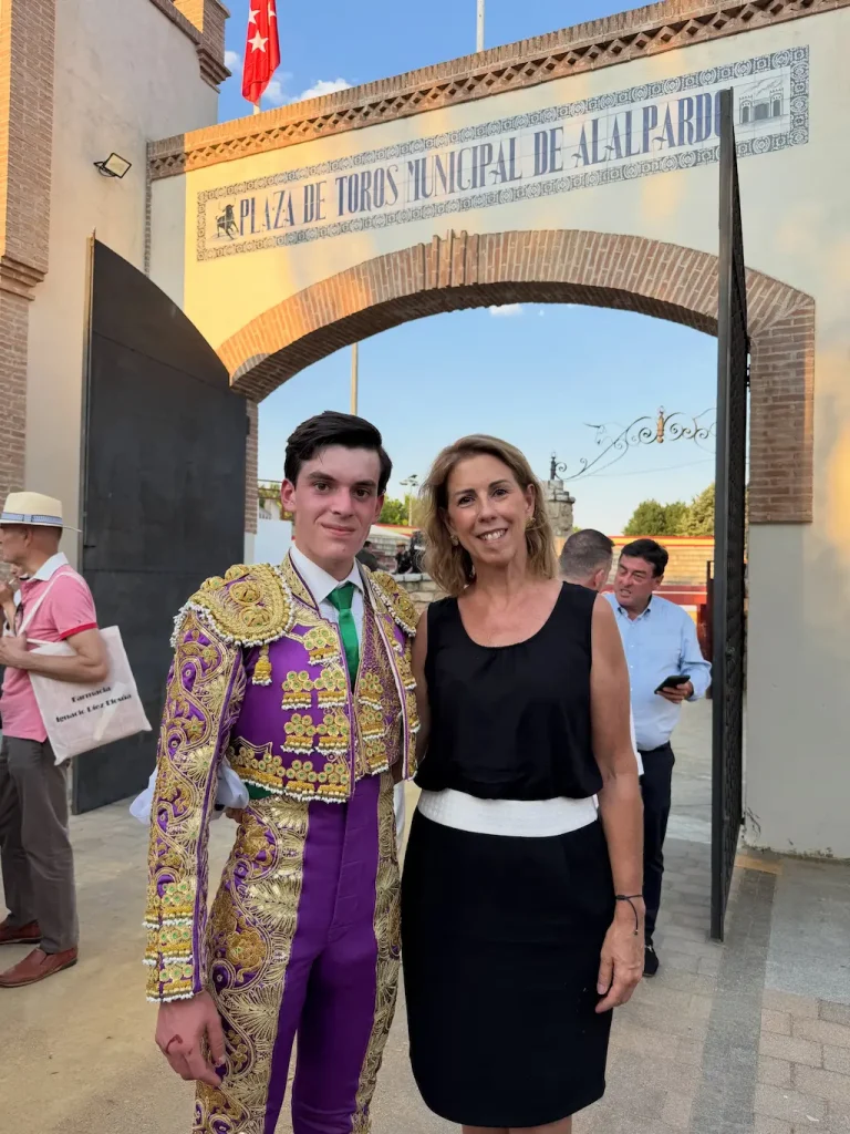 Sergio Rollón, vestido con un traje de torero morado y dorado, posa junto a una mujer vestida de blanco y negro a la entrada de la Plaza de Toros Municipal de Alalpardo durante el Circuito de Madrid. Al fondo se ve a otras personas.