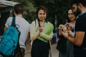 Cuatro jóvenes están al aire libre; tres de ellos disfrutan de unos helados en cucurucho, mientras que otro, con una mochila y una carpeta verde —quizá un «Borrador automático»—, se une a la conversación. Los frondosos árboles crean un agradable telón de fondo mientras disfrutan juntos de sus helados.