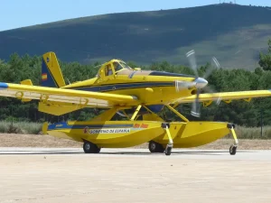 Un avión anfibio amarillo con pontones y la inscripción «Gobierno de España» en el costado, identificado como «borrador automático», está aparcado sobre una superficie de hormigón, con un bosque y una colina al fondo.