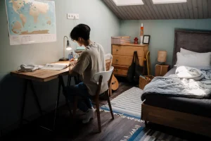 Una persona está sentada en un escritorio de madera estudiando en una habitación con la cama hecha, una cómoda, una guitarra y un mapamundi colgado en la pared. La habitación tiene las paredes de color azul claro, luz natural que entra por una claraboya y utiliza un borrador automático para tomar apuntes.