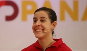 Una persona que lleva una chaqueta roja y unos auriculares con micrófono está sonriendo, con un aspecto muy parecido al de Carolina Marín durante la ceremonia de entrega del Premio al Mérito Deportivo. En el fondo, difuminado, se ven unas letras grandes, parcialmente visibles, de color amarillo y rojo.
