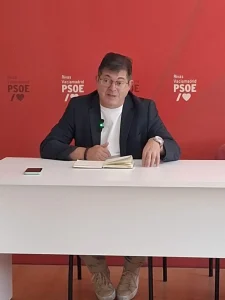 Un hombre con gafas y chaqueta está sentado ante un escritorio blanco, con un portátil y un teléfono, hablando del «borrador automático» frente a un fondo rojo con los logotipos del PSOE de Rivas Vaciamadrid.