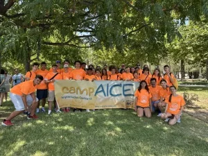 Un grupo de jóvenes, vestidos con camisetas naranjas a juego, están de pie al aire libre sobre el césped en Granada, sosteniendo una pancarta amarilla y azul en la que se lee «CAMP AICE - Implante Coclear». Se encuentran bajo unos árboles frondosos en un día soleado.