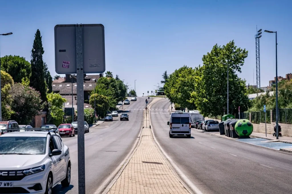 Una carretera ancha y desierta se extiende cuesta arriba bajo un cielo azul y despejado en la calle Junkal, con coches aparcados a ambos lados. Una furgoneta blanca circula por el centro, pasando junto a un carril bici. Árboles y casas bordean esta tranquila calle del barrio Oeste.