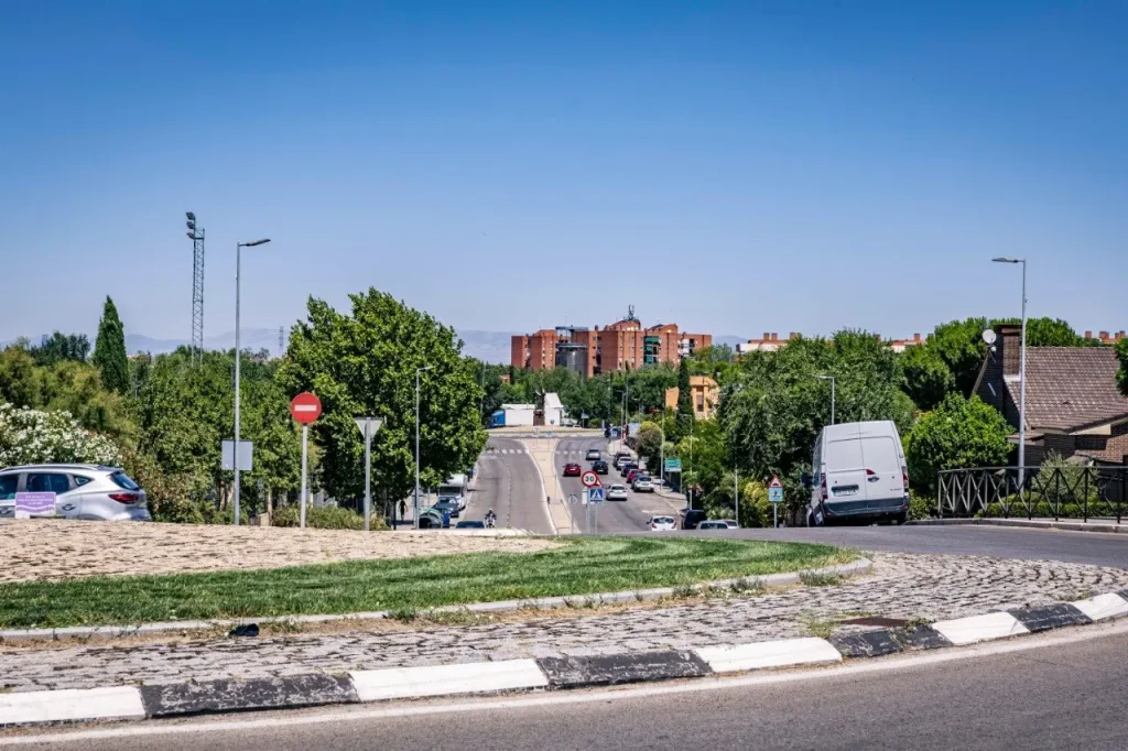 Una carretera asfaltada por la que circulan coches, una furgoneta blanca y un carril bici atraviesa el barrio Oeste por la calle Junkal, flanqueada por árboles, señales de tráfico y edificios residenciales bajo un cielo azul y despejado. Al fondo se alzan bloques de pisos de ladrillo rojo.