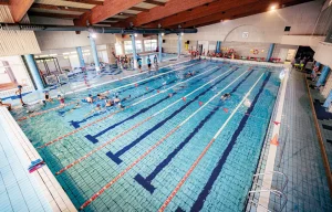 Piscina cubierta con varios carriles, nadadores en el agua y gente reunida en el extremo más alejado. La zona de la piscina cuenta con un suelo de baldosas, separadores de carriles rojos, un techo alto con vigas y una iluminación intensa. Se divisan algunos elementos del equipamiento de la piscina.