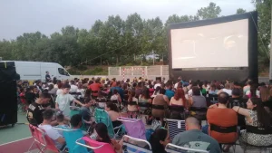 Un numeroso grupo de personas está sentado en sillas y bancos al aire libre en Rivas, disfrutando del cine de verano mientras ven películas gratis en una pantalla hinchable. Al fondo se ven árboles, y a un lado hay una furgoneta blanca aparcada.