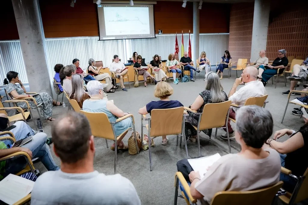 Un grupo de personas está sentado en círculo en una sala de reuniones de Rivas, escuchando a los ponentes que se encuentran al frente. En una pantalla de proyección se muestra un diagrama relacionado con un programa socioeducativo, con varias banderas y pancartas del Ayuntamiento de Rivas al fondo.